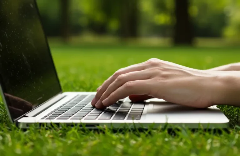 Mãos digitando em um notebook apoiado na grama, em ambiente ao ar livre, simbolizando a conexão entre tecnologia, mobilidade digital e sustentabilidade.